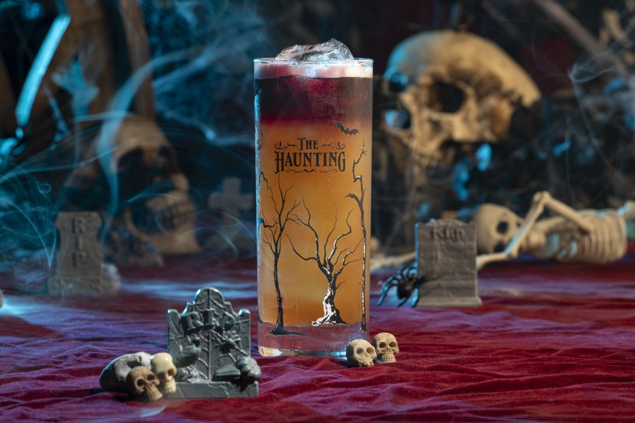 A layered red and orange collins cocktail beside a skull in blue fog.