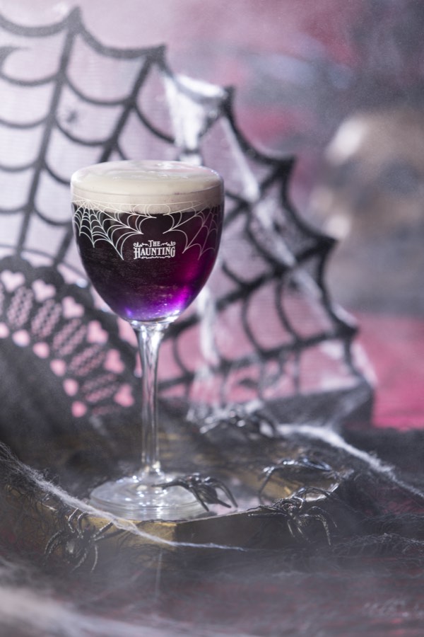 A deep purple cocktail with a pale foam cap in an etched Haunting coupe, set against black lace spider webs.