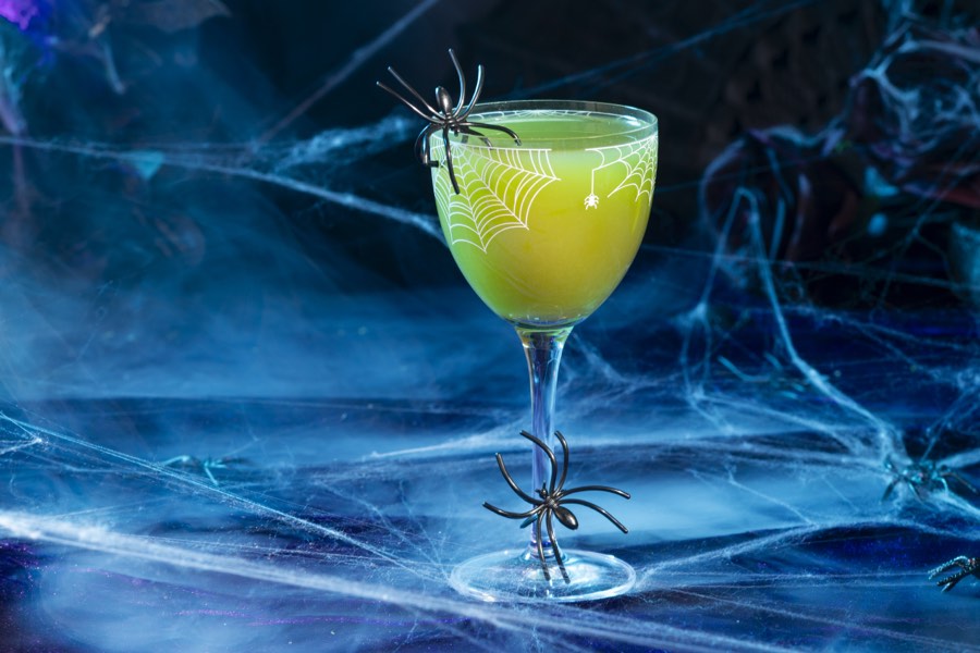 A chartreuse cocktail in a Nick and Nora glass behind glowing spider webs.